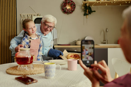 Caucasian senior man baking pie with granddaughter while grandmother taking photo using smartphone. Scene representing family bonding suitable for cooking campaigns