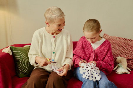 Senior Caucasian woman and child girl sitting together cutting paper snowflakes with scissors. Scene representing family bonding for holiday campaigns