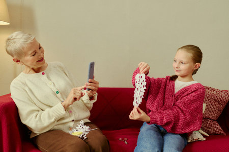 Senior Caucasian woman taking smartphone photo of Caucasian child showing paper craft snowflake. Image suitable for family lifestyle advertising and holiday crafting campaigns
