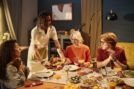 Young African American man serving roasted chicken to diverse young adult friends during dinner party. Concept representing holiday celebration, suitable for festive culinary