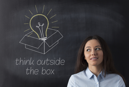 Young business woman standing with her back against a blackboard. She is looking to a big glowing light bulb.の写真素材