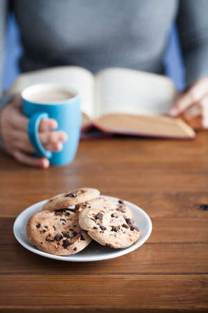Female hands holding hot drink mug and reading book over a table with chocolate chip cookies on saucerの写真素材