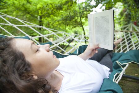 Woman lying in a hammock in a garden and enjoying a book readingの写真素材