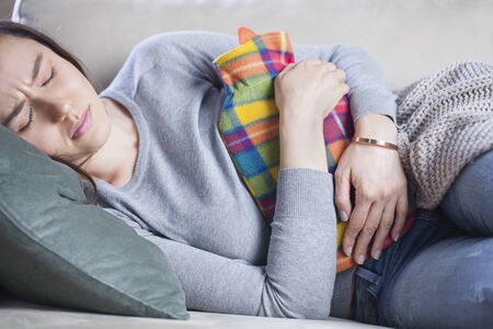 Young woman in painful expression, holding hot water bottle against belly suffering menstrual period pain lying down on sofa having tummy crampの写真素材