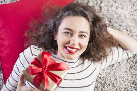 Happy pretty young woman holding gift box on carpetの写真素材
