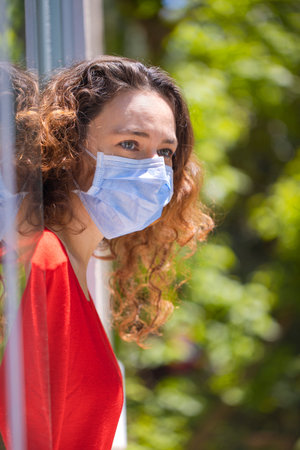 Young woman in medical mask on window breathing fresh air from quarantine