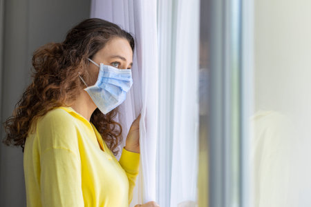 Young woman in isolation at home near window. Woman in quarantine looking through the window.