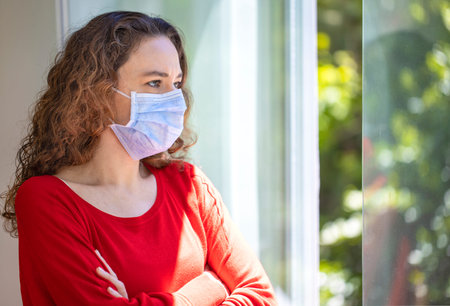 Young woman in medical mask on window breathing fresh air from quarantine