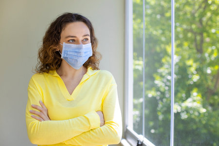 Young woman in medical mask on window breathing fresh air from quarantine