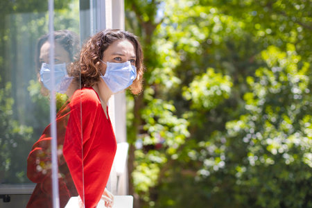 Young woman in medical mask on window breathing fresh air from quarantine