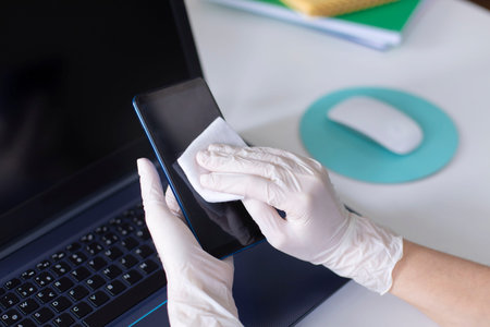 Woman hands with gloves wiping mobile phone while working from home. Protecting herself during  ...