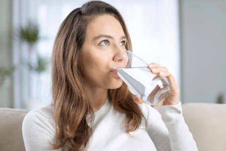 Young woman drinking water sitting on a couch at home. Health benefits of drinking enough water concepts.の写真素材
