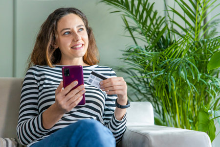 Smiling beautiful woman sitting on sofa and using smartphone while paying for online shopping with...