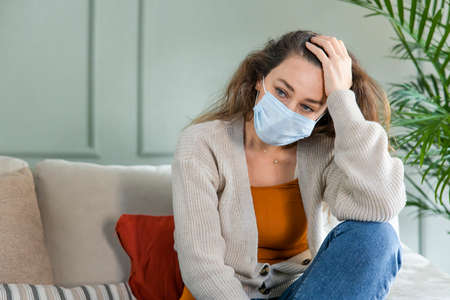 Sad young woman isolated stay at home in protective sterile mask on her face. Home quarantine to protect from pandemicの写真素材
