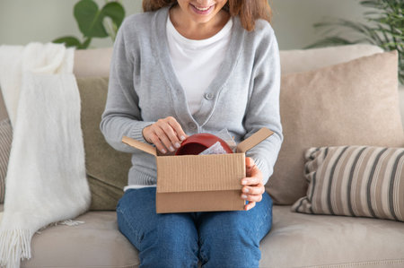 Young woman in her living room looks happy as she unpacks a small cardboard box containing something...