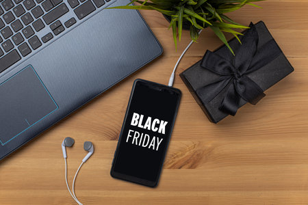 Black Friday shopping app in smart phone screen and black wrapped paper gift box on wooden table