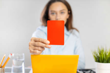 Young businesswoman holding red card and using laptop while sitting at the table. Dismissal or rejection conceptsの写真素材