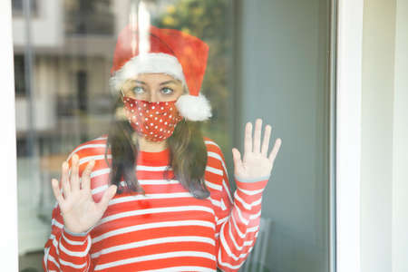 Young woman wearing a Santa hat and a protective face mask looking through a windowの写真素材