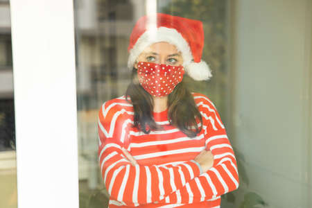 Young woman wearing a Santa hat and a protective face mask looking through a windowの写真素材