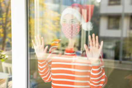 Young woman wearing a Santa hat and a protective face mask looking through a windowの写真素材