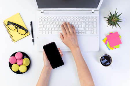 Female hands typing on laptop computer keyboard and holding smartphone. White office table, top view. Flat lay design.の写真素材