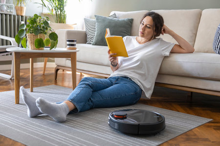 Robot vacuum cleaner cleaning the living room. Young woman enjoy rest, sitting on sofa at homeの写真素材