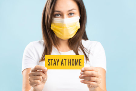 Young woman in face mask holding card with stay home lettering on isolated on blue background
