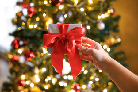 Woman hand with red ribbon gift box in front of Christmas tree