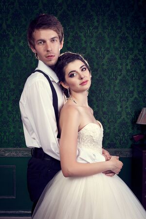 Happy bride and groom on green retro interior. Just married couple. Bride in wedding dress and groom is holding her from behind on green vintage wallの写真素材
