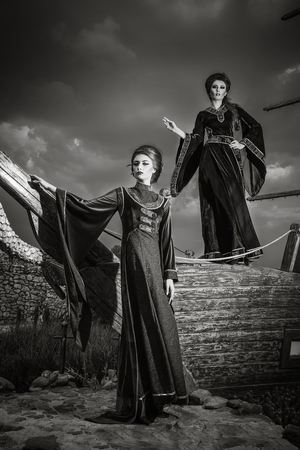 Two beautiful women dressed in vintage clothes in front of boat. Antique luxury. Retro clothes and vintage concept. Black and white imageの写真素材