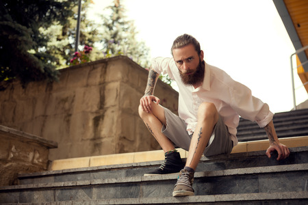 Beardead tattooed cool looking guy sitting in street on stairs looking away from the cameraの写真素材