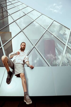 Hipster with long beard and tatoos in front of office building posing cool at cameraの写真素材