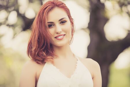 Beautiful redhead woman outdoor photo in summer on blurred tree flower backgroundの写真素材