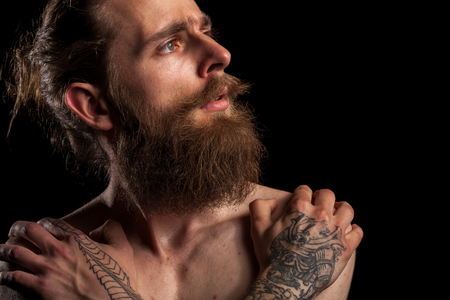 Portrait of tattooed bearded hipster guy on black background in studio photo. Expression and fashionの写真素材