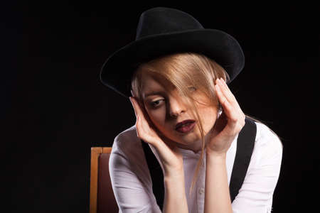 Blonde model with suspenders and white shirt wearing a hat on black background in studio photo