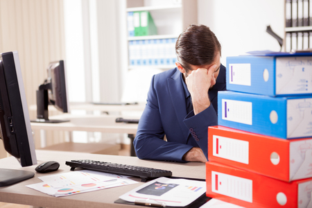 Corporate worker depressed from the workload at the office. His desk is full with a pile of folders and papers with finance charts are on the table.の写真素材