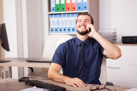 Smiling CEO in blue shirt and tie talking on the phone. Portrait of successful entrepreneur in his modern office. Happy young corporate worker at his jobの写真素材