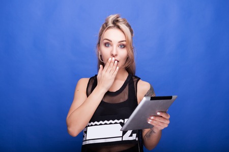 Surprised young woman holding her hand at the mouth and in the other hand holding a digital tablet. Studio photoの写真素材