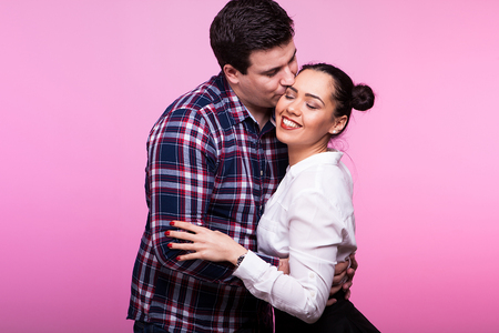 Happy husband kissing his wife on the cheek in studio on pink backgroundの写真素材
