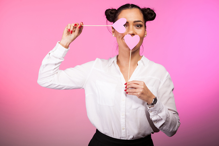 Beautiful woman covering her eye and mouth with pink hearts while looking at the camera. Pink background and studio photoの写真素材