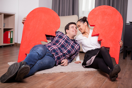 Happy couple holding two parts of one big heart lying on the floorの写真素材
