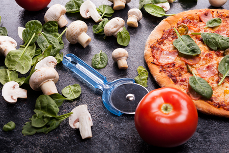 Homemade pizza next to mushrooms, baby spinach and tomatoes with a pizza cutter on dark wooden backgroundの写真素材
