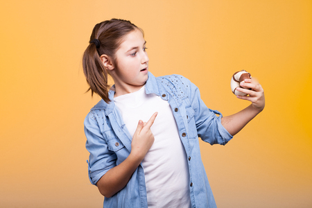 Expressive young girl poiting at a donut she is holding in hands on yellow backgroundの写真素材