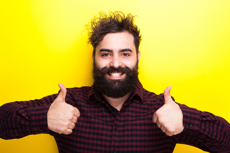 Smiling bearded hipster with his thumbs up on yellow backgroundの写真素材