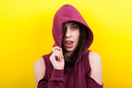 Hipster woman wearing a hood and winking to the camera on yellow background in studioの写真素材