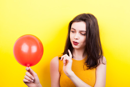 Smiling young woman playing with a red balloon on yellow background in studioの写真素材