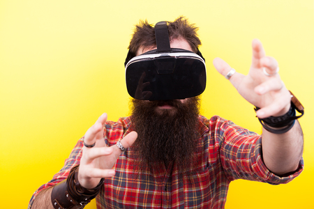 Long bearded hipster man wearing a virtual reality VR headset and makes gestures on yellow backgroundの写真素材