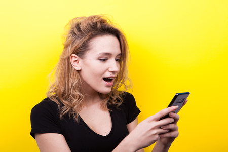 Surprised woman with a phone in hands on yellow background in studio photoの写真素材