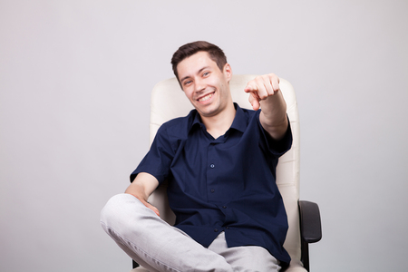 Happy smiling successful young businessman in casual blue shirt sitting on an office chair over gray background in studio photoの写真素材