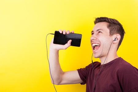 Joyful man with headphones in ears singing in the phone like it is a microphone. Yellow backgorund....